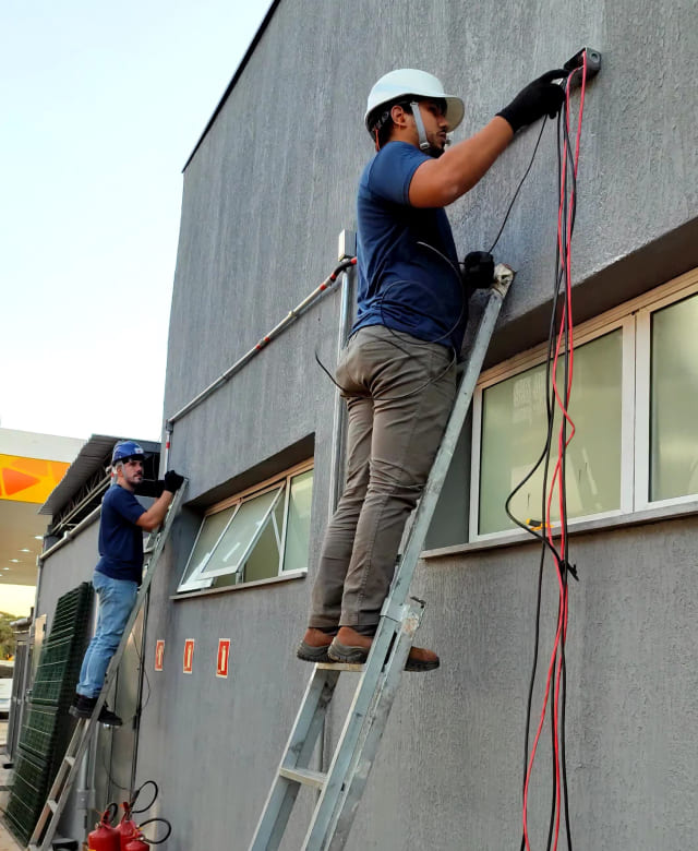 Dois técnicos da Enlight Engenharia realizam a instalação do sistema de alarme de incêndio na fachada externa de um posto de combustíveis, com uso de escadas, EPIs e cabeamento técnico.