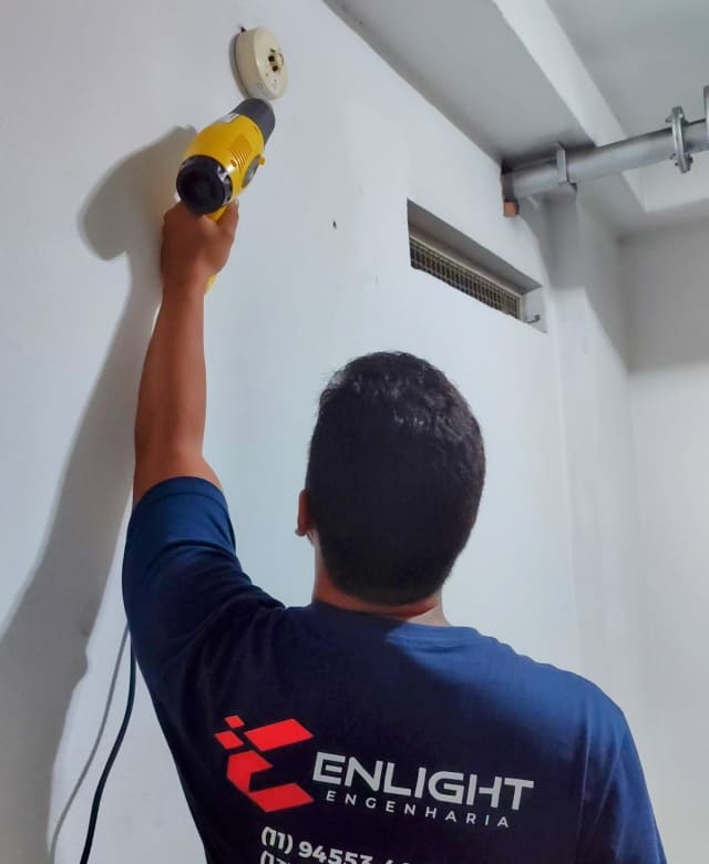Técnico da Enlight Engenharia aplica calor com soprador térmico em detector de incêndio instalado na parede