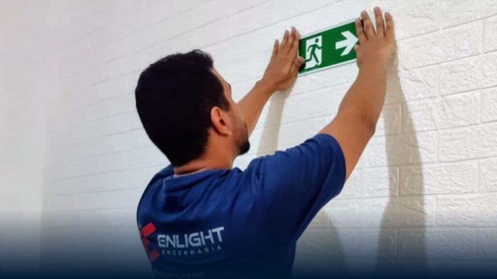 Profissional instalando placa de sinalização de emergência fotoluminescente de saída, demonstrando adequação à IT-20/2025 em edificação da Baixada Santista.