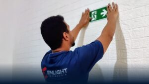 Profissional instalando placa de sinalização de emergência fotoluminescente de saída, demonstrando adequação à IT-20/2025 em edificação da Baixada Santista.