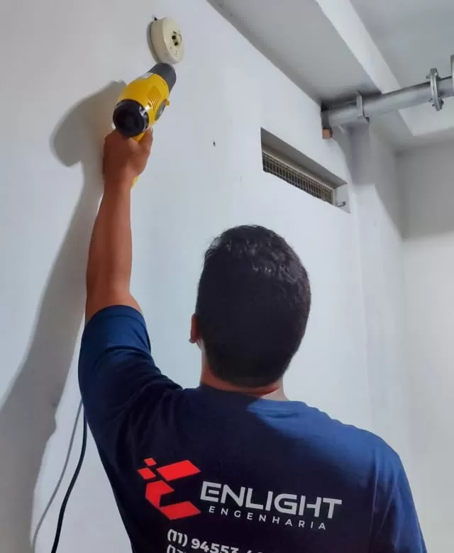 Técnico da Enlight Engenharia aplica calor com soprador térmico em detector de incêndio instalado na parede