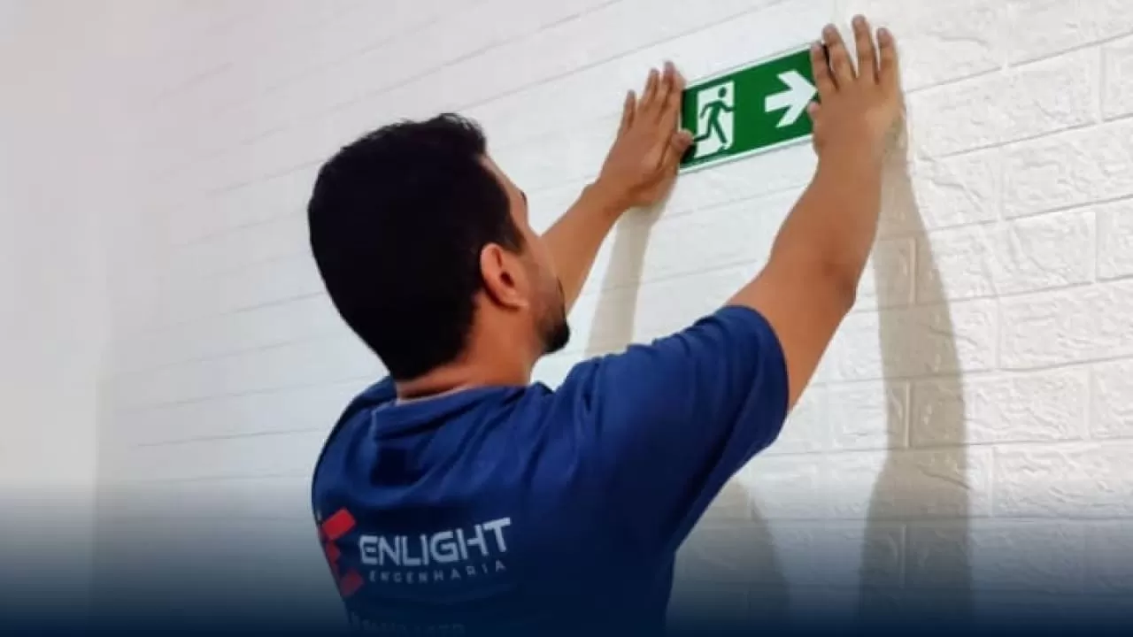 Profissional instalando placa de sinalização de emergência fotoluminescente de saída, demonstrando adequação à IT-20/2025 em edificação da Baixada Santista.